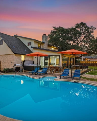 Backyard with illuminated stone and metal house, curved swimming pool, blue lounge chairs, orange umbrellas, grill, and volleyball net at dusk