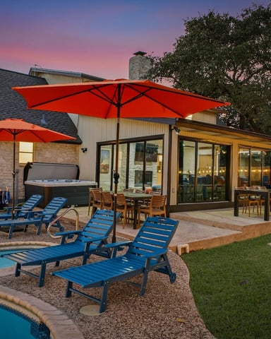 Backyard patio at dusk with swimming pool, blue lounge chairs, red umbrellas, and modern house with glass doors