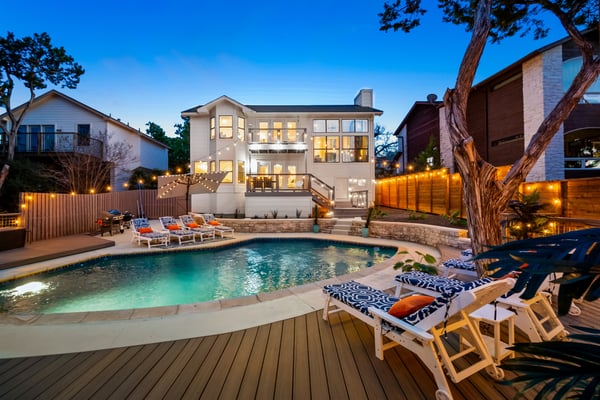 Aerial view of modern home with illuminated pool, hot tub, lounge chairs, and patio at dusk overlooking neighborhood